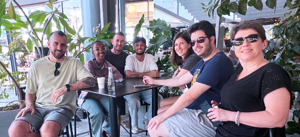 Playtika design team — seven colleagues gathered around a table on a sunlit terrace with tropical plants